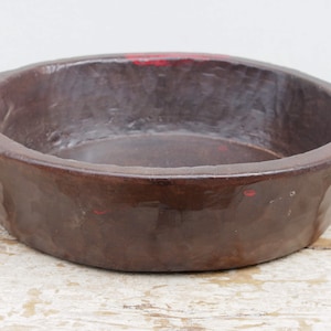 Può includere: Una ciotola rotonda in legno marrone scuro con un bordo largo. La ciotola ha una finitura liscia e lucida con venature del legno visibili. Piccoli segni di vernice rossa sono visibili sul bordo. La ciotola poggia su una superficie di legno chiaro e invecchiato.