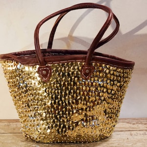 Puede incluir: Un bolso tote de lentejuelas doradas con asas de cuero marrón. El bolso está hecho de un material tejido y tiene una forma redonda.