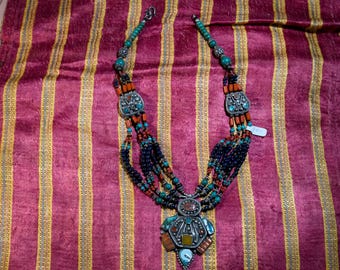 May include: A multi-strand necklace with turquoise, coral, and black beads. The necklace has a large silver pendant with turquoise and coral accents. The necklace is displayed on a striped fabric background.