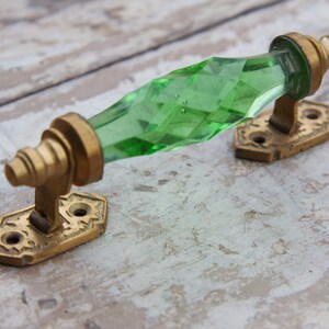 May include: A vintage door pull with a faceted, emerald-green glass handle. The handle is mounted on aged brass hardware with decorative detailing. The hardware includes two mounting plates with screw holes for installation. The overall design is ornate and antique.