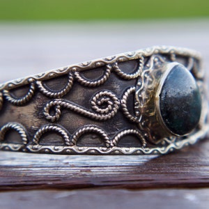 Puede incluir: Una pulsera de puño plateada con un diseño ornamentado. Presenta una piedra grande, ovalada, de color verde oscuro engastada en un bisel decorativo. El trabajo en metal incluye patrones de cuerda y volutas, creando una estética vintage o bohemia.