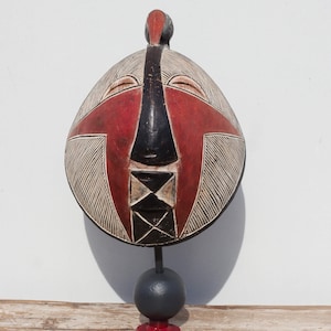 May include: A wooden mask with a red, black and white geometric design. The mask is on a stand and has a bird-like head.