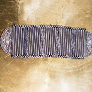 Puede incluir: Un brazalete de plata con un diseño decorativo. El brazalete está hecho de muchas piezas pequeñas unidas que se pueden ajustar para adaptarse a diferentes tamaños de muñeca.