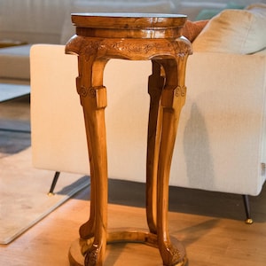 Peut inclure: Un support de plante en bois avec un dessus et une base ronds, et trois pieds incurvés. Le support est en bois brun clair et présente des détails floraux sculptés sur les pieds et le dessus. Le support mesure environ 71 cm de haut.