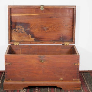 Puede incluir: Un cofre de almacenamiento de madera con bisagras de latón y un pestillo de latón. El cofre está abierto y el interior es visible. El cofre está sentado sobre una alfombra estampada.