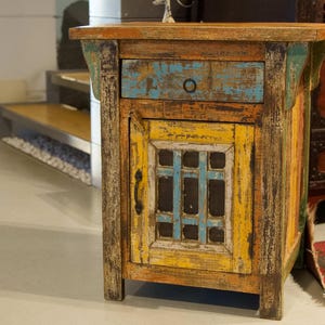 May include: A small, rustic wooden cabinet with a distressed finish. The cabinet features a drawer with a blue painted front and a door with a yellow and blue grid design. The wood has orange, blue, and yellow tones, giving it a vintage look.