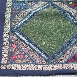 May include: A square throw pillow with a black background and a diamond-shaped green center. The diamond is surrounded by a black and silver trim. The pillow is made of fabric and has a variety of colorful embroidered details.