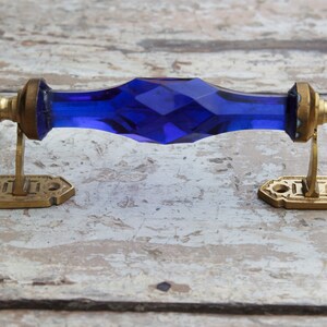 Op de afbeelding: Een vintage deurklink met een facetgeslepen, diepblauwe glazen middenstuk en messing hardware. De klink heeft een rechthoekige vorm met decoratieve messing platen aan elk uiteinde. Het messing heeft een verouderde uitstraling.