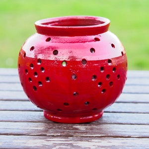 Peut inclure: Une lanterne en céramique rouge de forme ronde avec de nombreux petits trous. La lanterne est posée sur une surface en bois.
