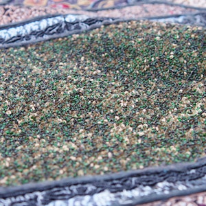 May include: Close-up of a fabric with a textured surface. The fabric is made of small, green, brown, and black sequins. The sequins are arranged in a random pattern.