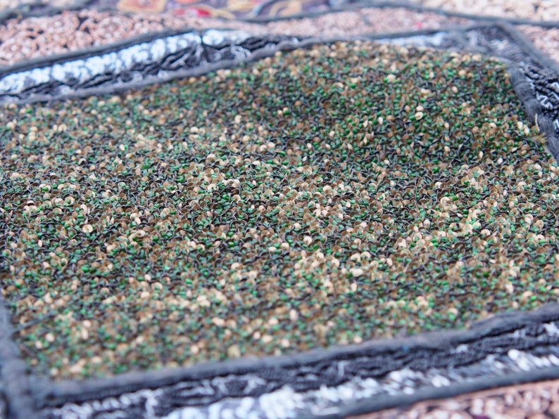 May include: Close-up of a fabric with a textured surface. The fabric is made of small, green, brown, and black sequins. The sequins are arranged in a random pattern.