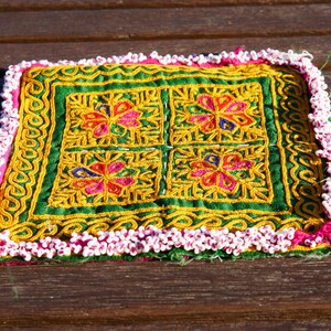 Könnte beinhalten: Ein quadratisches grünes Stoffkissen mit einem weißen Perlenrand. Das Kissen ist mit vier gestickten Blumenmotiven in den Farben Rosa, Gelb, Blau und Grün verziert.