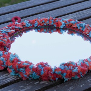 May include: A round mirror with a blue and orange fringe border. The mirror has a red fabric loop for hanging.