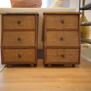 Op de afbeelding: Twee bijpassende houten nachtkastjes met elk drie lades. De lades hebben een getextureerde, lichtbruine voorkant en een donkerbruin frame. Elke lade heeft een ronde, donkere metalen handgreep. De nachtkastjes staan op korte, afgeronde poten.