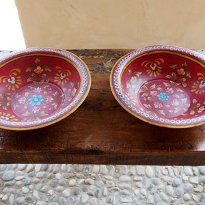 Puede incluir: Dos cuencos rojos con intrincados diseños florales pintados en oro, azul y rosa. Los cuencos están sobre una mesa de madera.