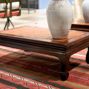Puede incluir: Una mesa de centro marrón oscuro con una parte superior tejida y patas curvas. Un jarrón de cerámica blanca está sobre la mesa. La mesa está sobre una alfombra estampada con rayas rojas, negras y blancas.