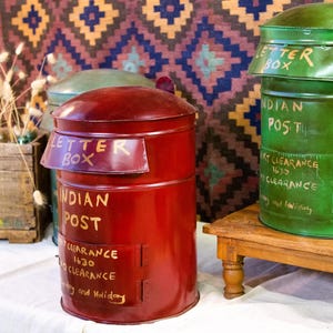 Op de afbeelding: Drie vintage Indiase postbrievenbussen. De rode brievenbus staat op de voorgrond, met de tekst "LETTER BOX" en "INDIAN POST" in gouden letters. Een groene brievenbus staat op een houten standaard. De achtergrond is een textiel met patronen.