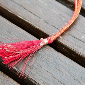 May include: A red and gold cord with a red tassel. The cord is lying on a wooden surface.