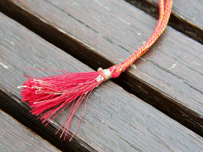 May include: A red and gold cord with a red tassel. The cord is lying on a wooden surface.