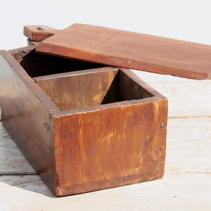 Può includere: Una scatola di legno vintage con un coperchio incernierato e due scomparti. La scatola è in legno marrone scuro e ha un aspetto rustico.