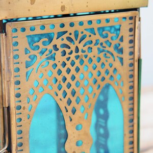 May include: A brass lantern with a teal blue glass panel and intricate cut-out designs. The lantern has a square shape and a hinged door.