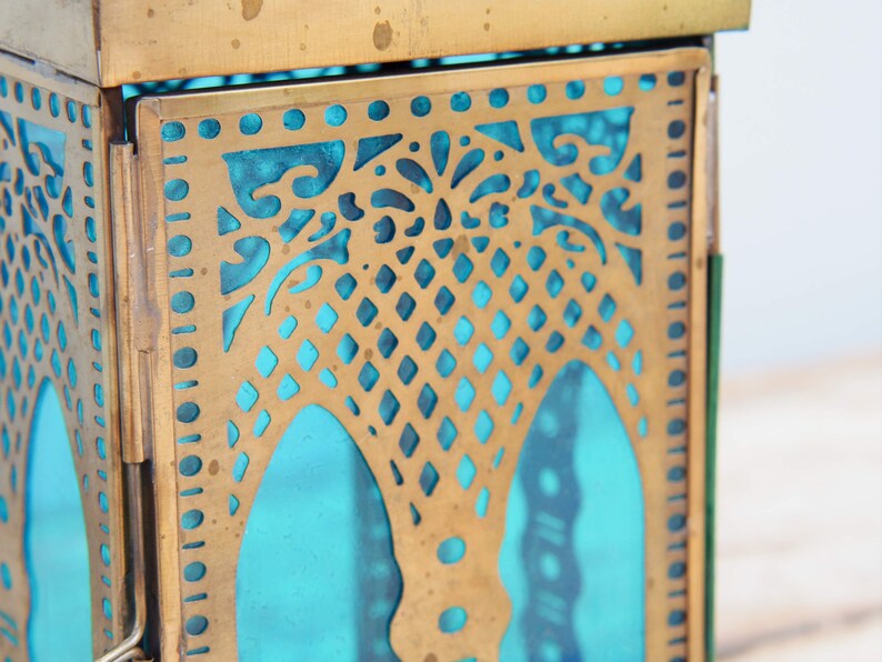 May include: A brass lantern with a teal blue glass panel and intricate cut-out designs. The lantern has a square shape and a hinged door.