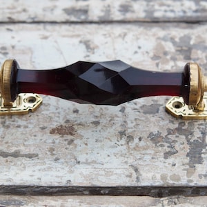 May include: A decorative cabinet pull with a faceted, dark red center and brass-colored end pieces and mounting plates. The pull is designed with a geometric shape and is mounted on a weathered, light-colored surface.