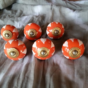 Puede incluir: Ocho pomos de cerámica naranja y blanco con un acento dorado. Los pomos tienen un diseño floral.