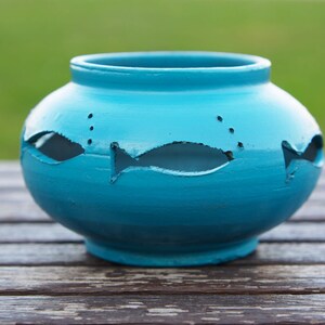 Può includere: Un vaso in ceramica turchese con intagli a forma di pesce. Il vaso ha un corpo arrotondato e un bordo leggermente svasato. Gli intagli a forma di pesce sono distribuiti uniformemente attorno al vaso, con piccoli intagli circolari sopra. Il vaso è su una superficie di legno.