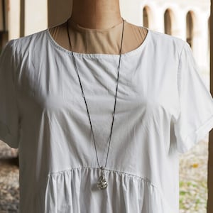 Può includere: Un abito bianco in cotone con una collana di cordoncino nero e un ciondolo d'argento.