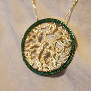 Może przedstawiać: Złoty naszyjnik z okrągłym, zielono-białym wisiorem. Wisiór przedstawia drzewo życia z hebrajskimi literami.