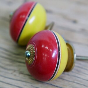 Può includere: Due pomelli rotondi in legno per armadio con un design di vernice rosso e giallo. I pomelli hanno una striscia nera e una vite in metallo dorato.