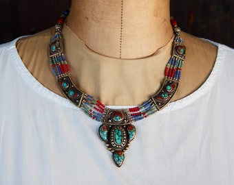 May include: A multi-strand necklace with a large turquoise and coral pendant. The necklace is made of silver and features blue and red beads.