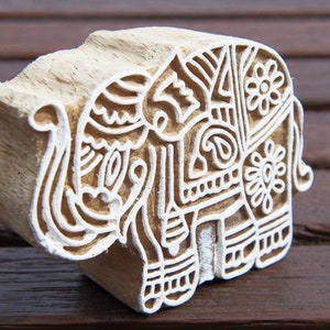 Op de afbeelding: Een houten bloksnijwerk van een olifant met ingewikkelde witte ontwerpen. De olifant kijkt naar rechts en heeft zijn slurf omhoog gestoken.