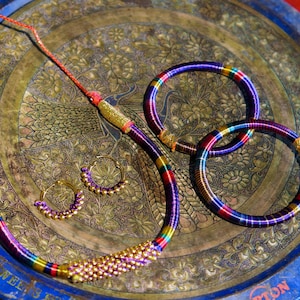 Op de afbeelding: Een kleurrijke sieradenset met een ketting, oorbellen en twee armbanden. De ketting en armbanden zijn gemaakt van een geweven materiaal met regenboogstrepen. De oorbellen zijn goudkleurige creolen met paarse en gouden accenten. De set wordt gepresenteerd op een decoratief dienblad.