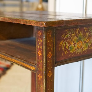 May include: A small, antique wooden side table with a dark reddish-brown finish. The table features a lower shelf and is decorated with floral designs in shades of green, yellow, and orange. The edges are adorned with small, repeating floral patterns.