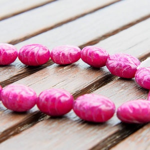 May include: A strand of pink oval-shaped beads with a carved design. The beads are strung on a white cord and are lying on a wooden surface.