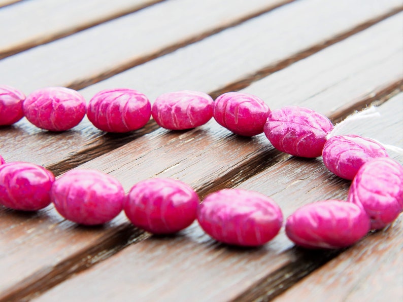 May include: A strand of pink oval-shaped beads with a carved design. The beads are strung on a white cord and are lying on a wooden surface.