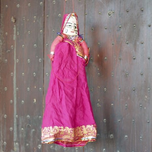 Può includere: Un burattino tradizionale indiano con un vestito di stoffa rosa e un viso dipinto. Il burattino è appeso a una corda su uno sfondo di legno.