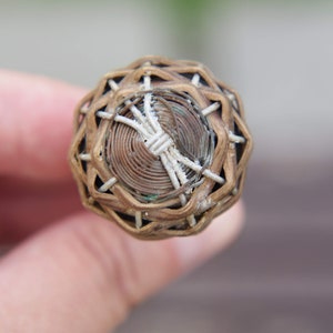 Puede incluir: Un primer plano de una cesta pequeña, redonda y tejida con un patrón marrón y blanco. La cesta está hecha de materiales naturales y tiene un diseño delicado e intrincado.