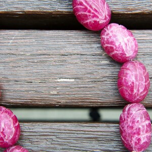 May include: A close-up of a pink carved stone necklace on a wooden surface. The necklace is made of oval-shaped beads with a carved leaf design.