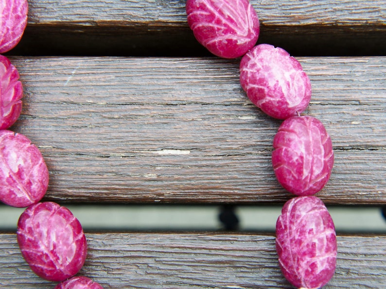 May include: A close-up of a pink carved stone necklace on a wooden surface. The necklace is made of oval-shaped beads with a carved leaf design.