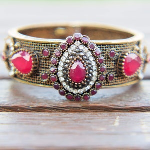 Peut inclure: Un bracelet jonc en or avec un motif décoratif composé de pierres précieuses rouges et transparentes. Le bracelet présente un motif tissé et est orné de petites pierres rouges.