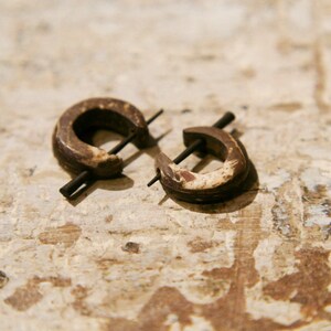 Può includere: Due orecchini a cerchio in legno marrone con un perno di metallo al centro. Gli orecchini sono su una superficie in legno marrone chiaro.