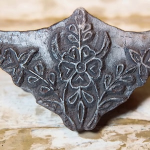 Pode incluir: Um carimbo de impressão de bloco de metal cinza vintage com um design floral. O design apresenta uma flor grande com uma flor menor de cada lado, cercada por folhas.