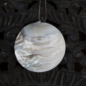 Może przedstawiać: Biały, opalizujący, okrągły wisiorek zawieszony na srebrnym łańcuszku. Wisiorek wykonany jest z perłowej muszli i ma naturalny, organiczny wzór.