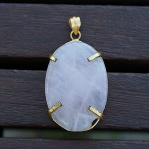 Może przedstawiać: Owalny wisiorek z jasnoróżowego kamienia szlachetnego z złotą oprawą. Wisiorek jest osadzony w prostym, eleganckim wzorze z czterema złotymi szponami, które trzymają kamień.