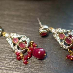 May include: A pair of ornate earrings with ruby-coloured gemstones and small red beads. The earrings have a silver-coloured metal setting with clear accent stones and gold-coloured accents.