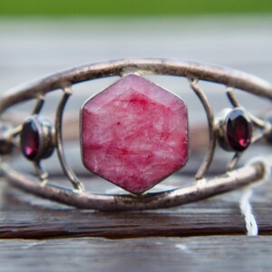 May include: A silver-toned bracelet featuring a large, faceted, pink gemstone at its center, flanked by smaller, round, dark red gemstones. The bracelet has an open design with decorative metalwork.