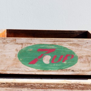 Può includere: Una cassa di legno vintage dipinta di bianco con un logo "7 Up" verde e rosso. La cassa ha un aspetto usurato e rustico.